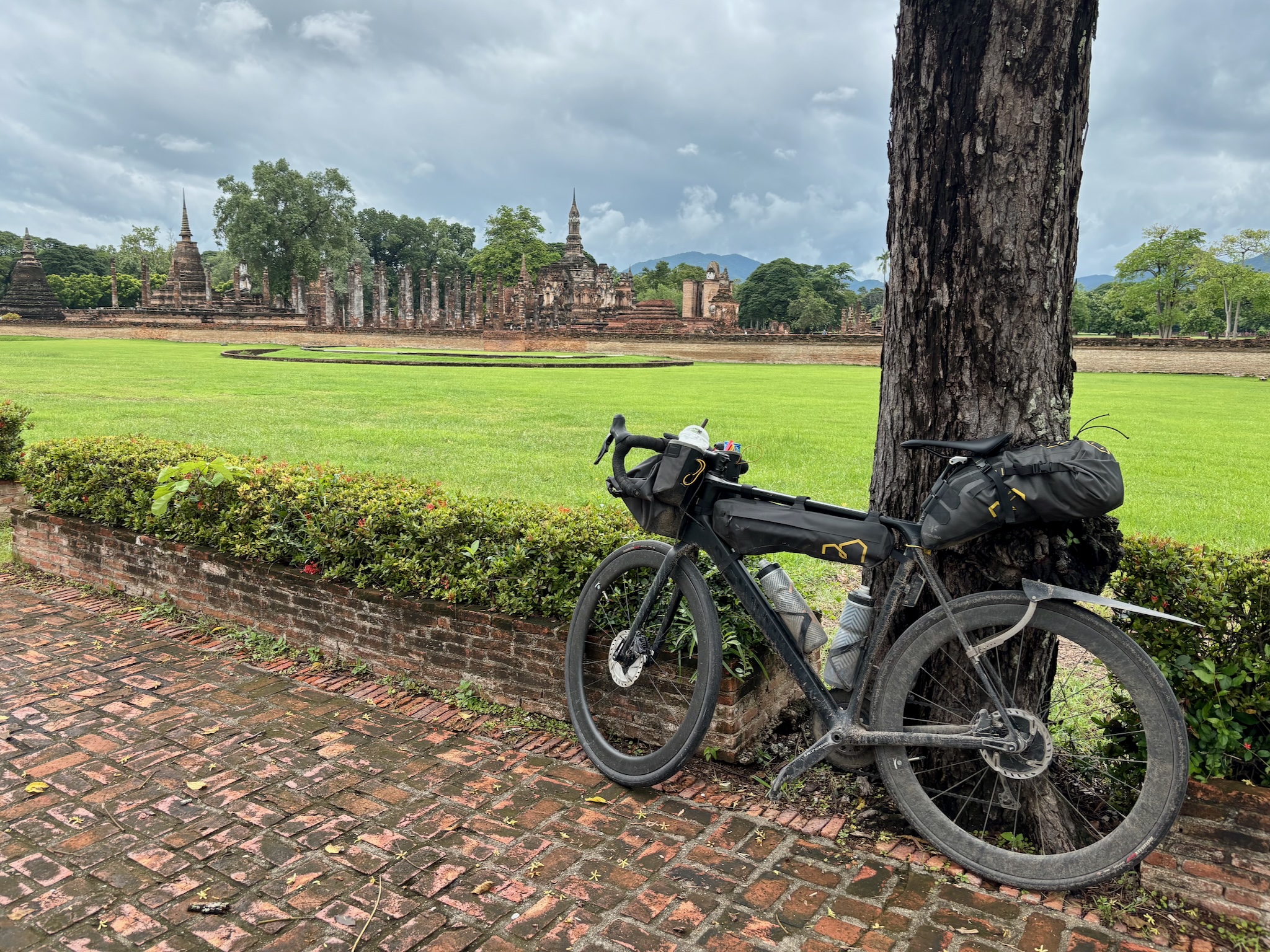 The loaded bike leaned against a tree at Sukhothai historical park, ruins across the lawn