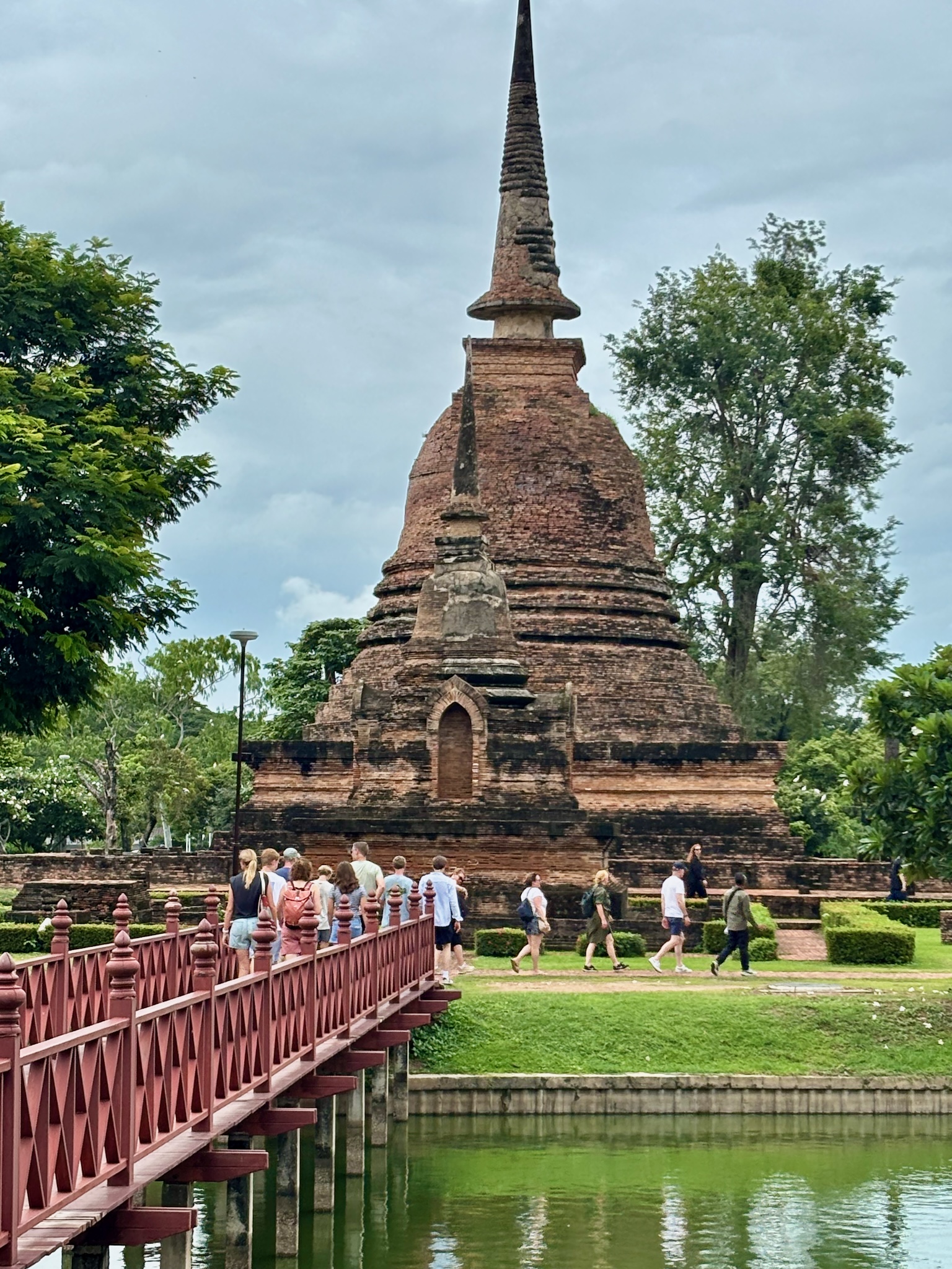 A brick chedi with its spire, visitors crossing a red bridge to reach it