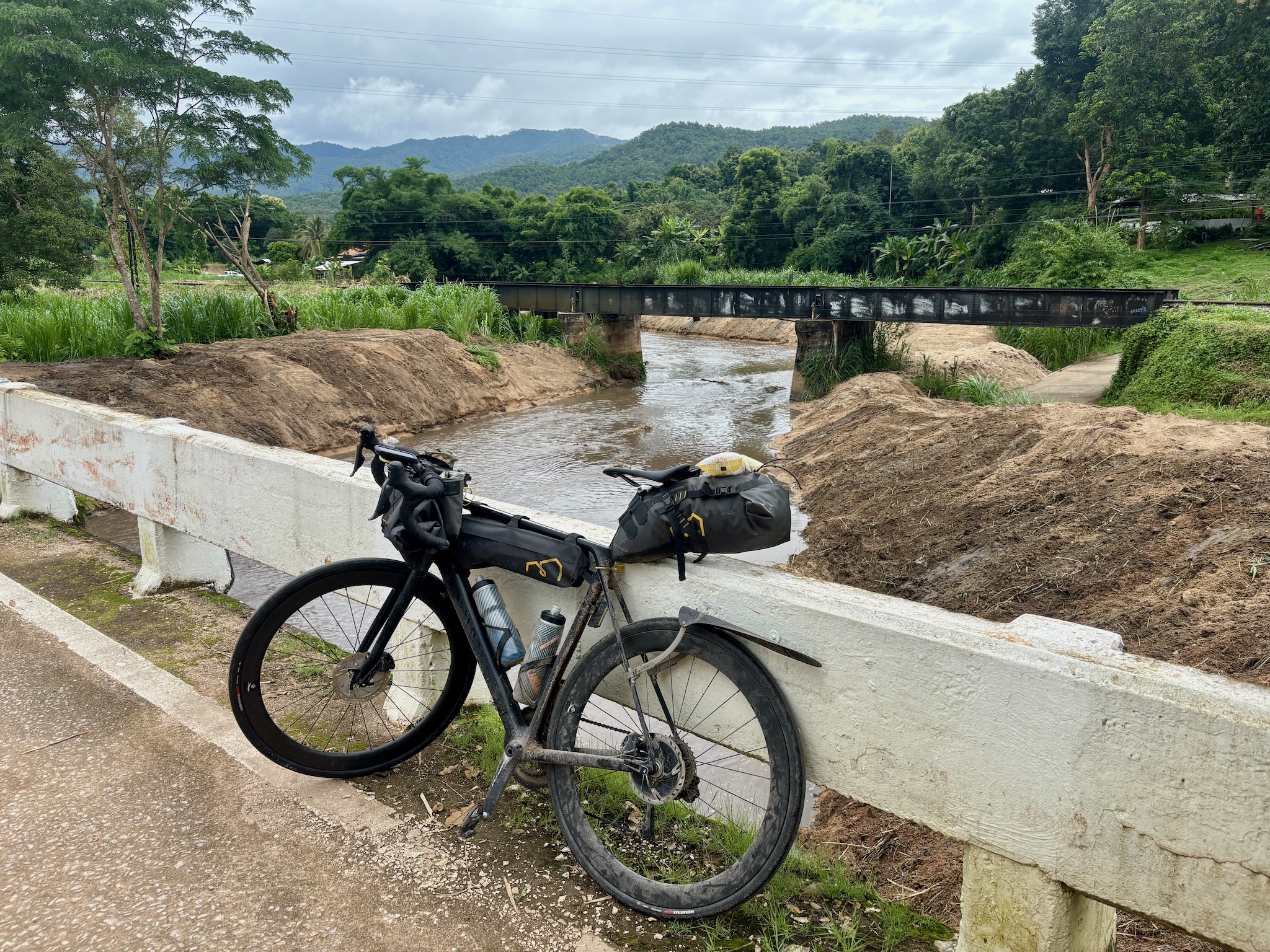The loaded bike on a concrete bridge over a muddy river, a railway bridge and green hills beyond