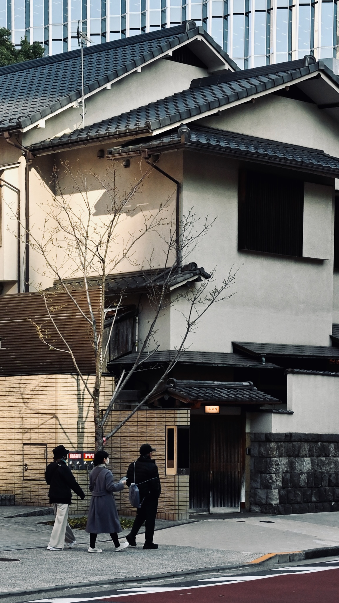 A traditional tiled house below a glass office tower, people walking past
