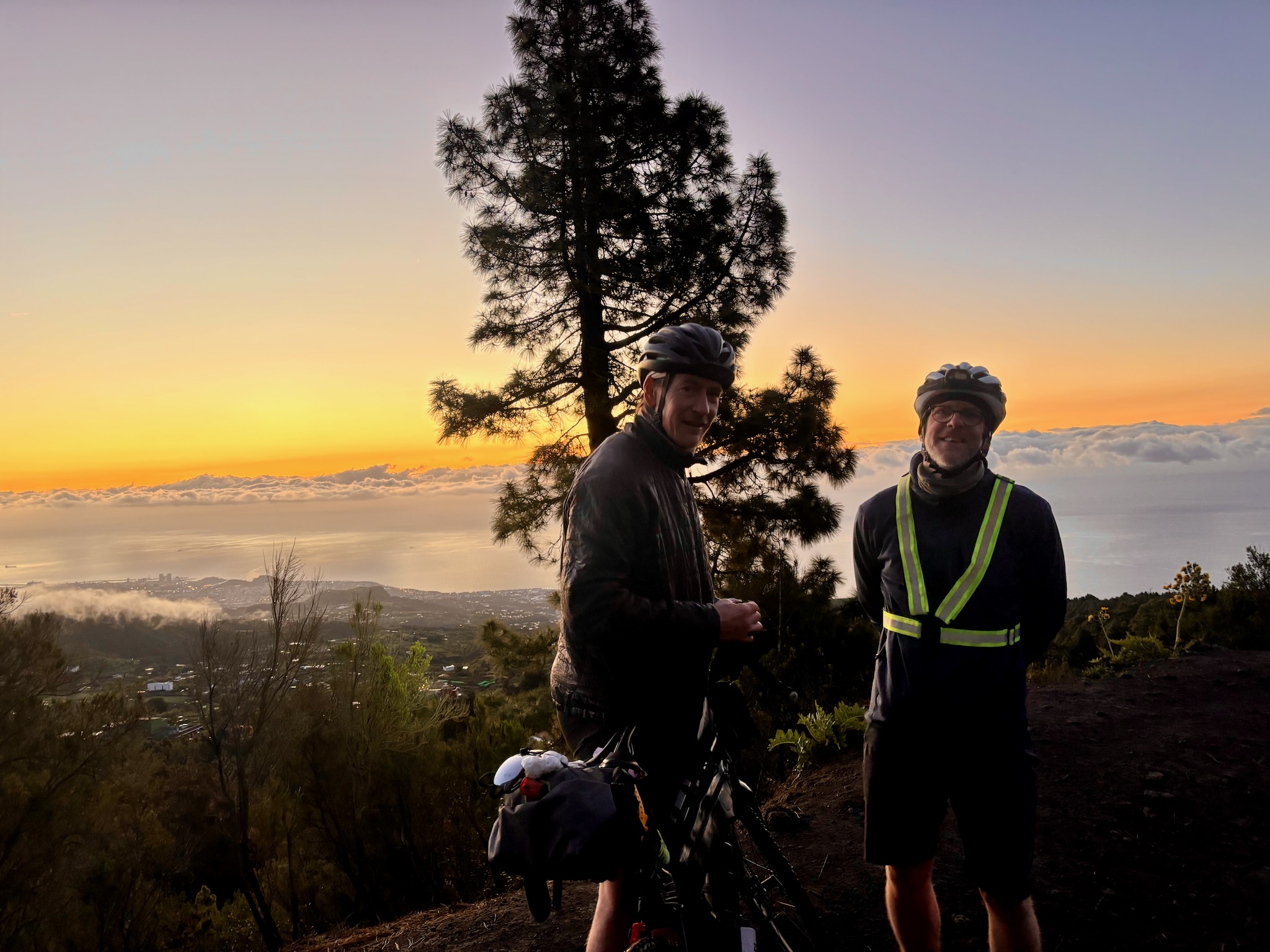 Two riders at a pine-topped viewpoint above a sea of cloud at sunset