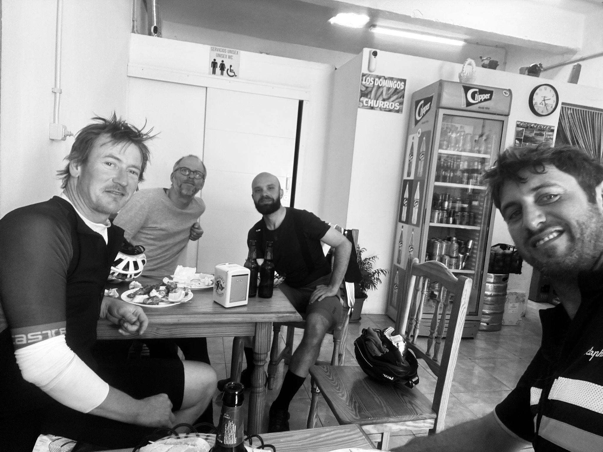 Four riders sharing beer and food around a table at a roadside bar, in black and white