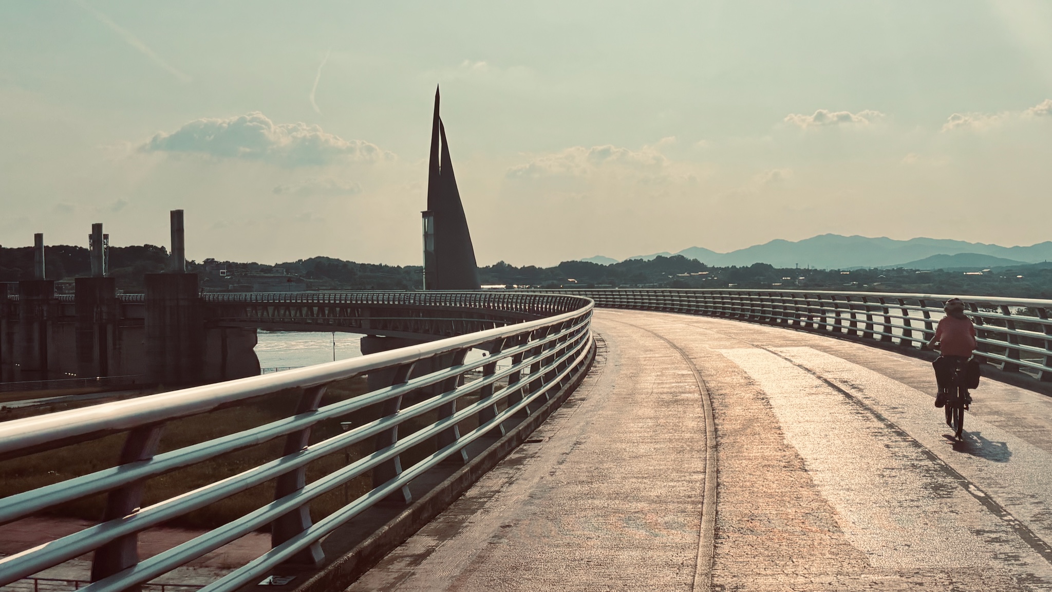 A cyclist crossing a curved bridge beside a sail-shaped weir tower